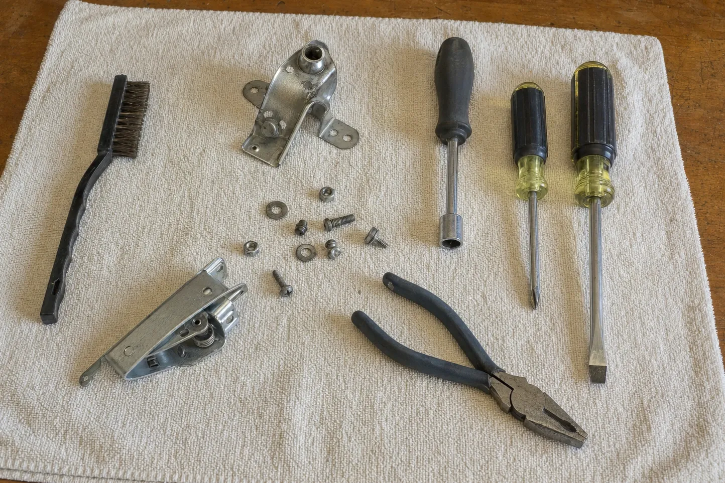 Wire brush, a cast bracket with a hollow boss, and a latch assembly with its spring and cam laid out on a towel with a nut driver, two screwdrivers and lineman’s pliers